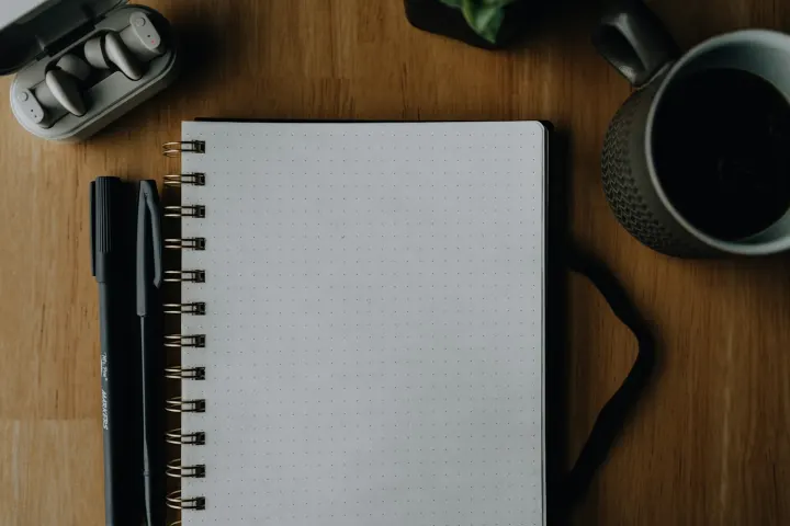 Ahşap bir masada yazılmayı bekleyen noktalı defter ve siyah kalem.
