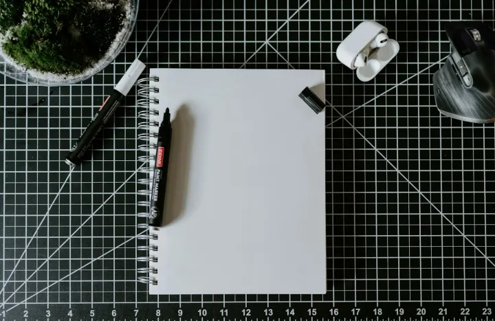 An open notebook, pens, mouse and earbuds arranged on a gridded desk mat.